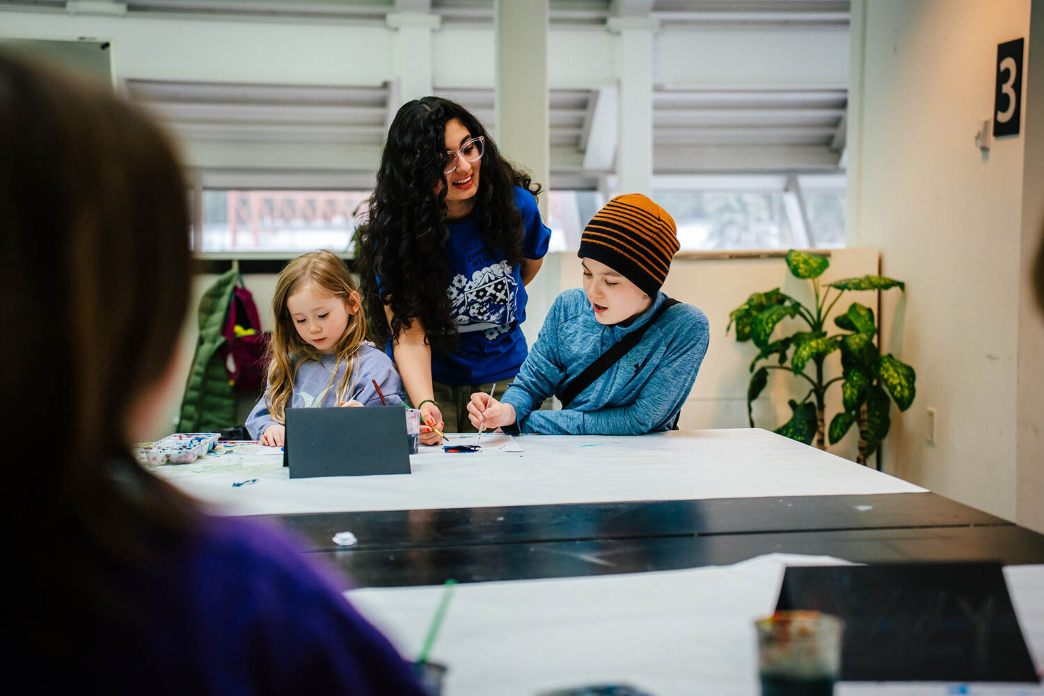 An older youth helps two younger children with a painting at a table.