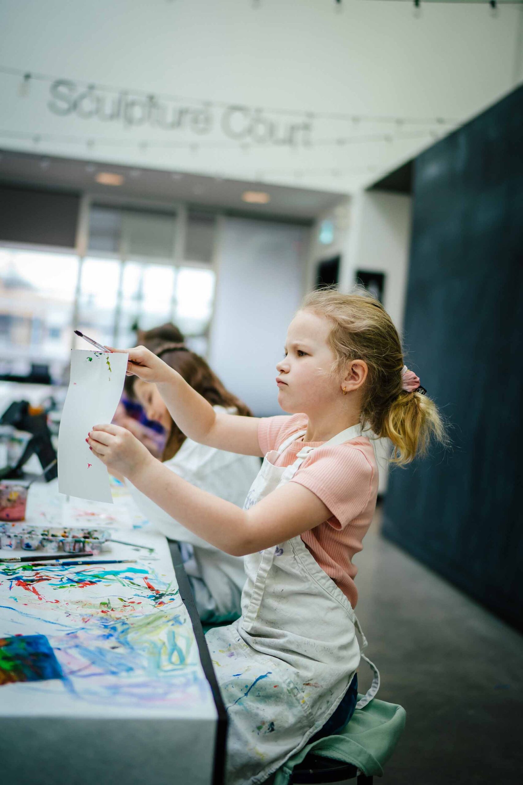 A child who is very focused on their painting, holding up a paper and brush in the air above a very messy table covered in paint.