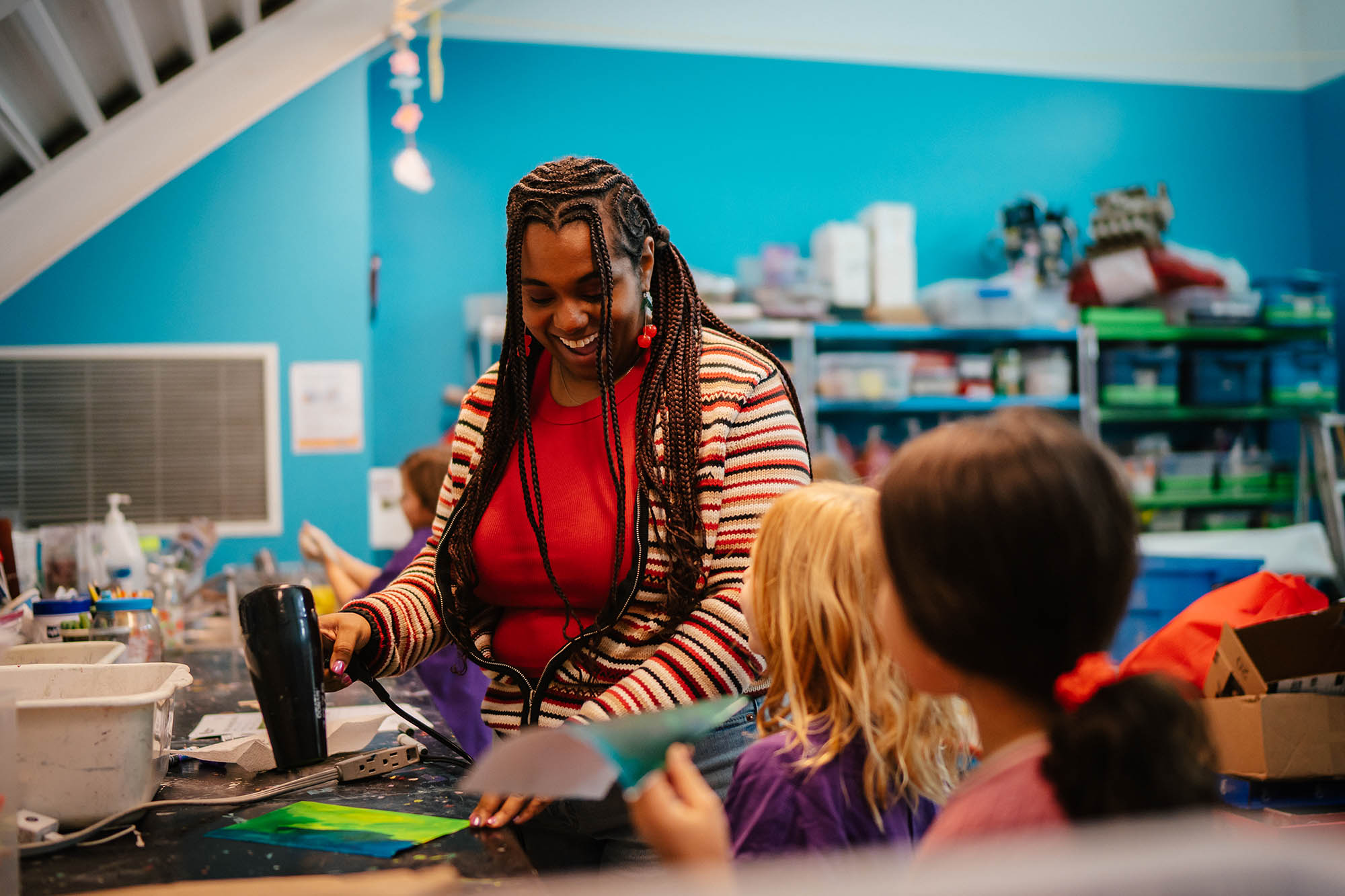 An adult uses a hairdryer to dry a painting for two children waiting in front of her, they are all laughing and smiling big.